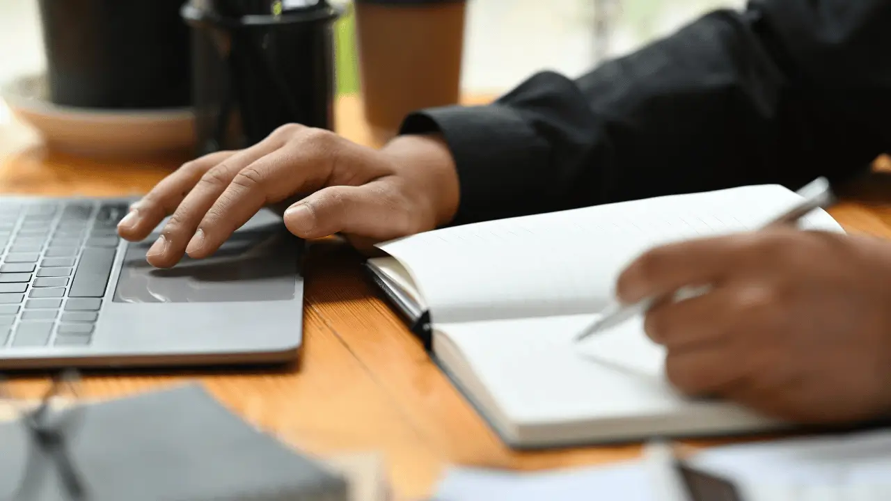 Homem mexendo no notebook e ao mesmo tempo escrevendo em uma agenda. Ele é branco e usa roupa social.