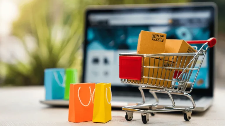 Mini carrinho de compras com sacolas e um notebook desfocado atrás.