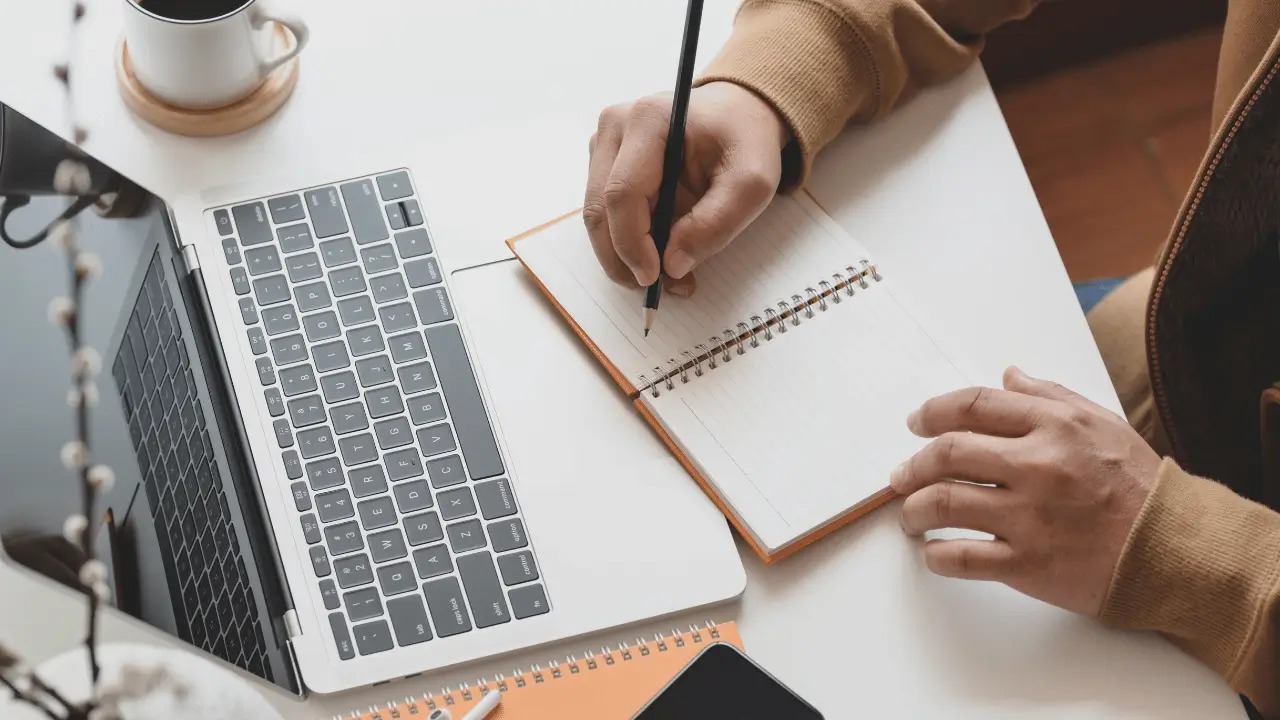 Imagem mostra homem escrevendo em um caderno e com um notebook aberto em sua frente