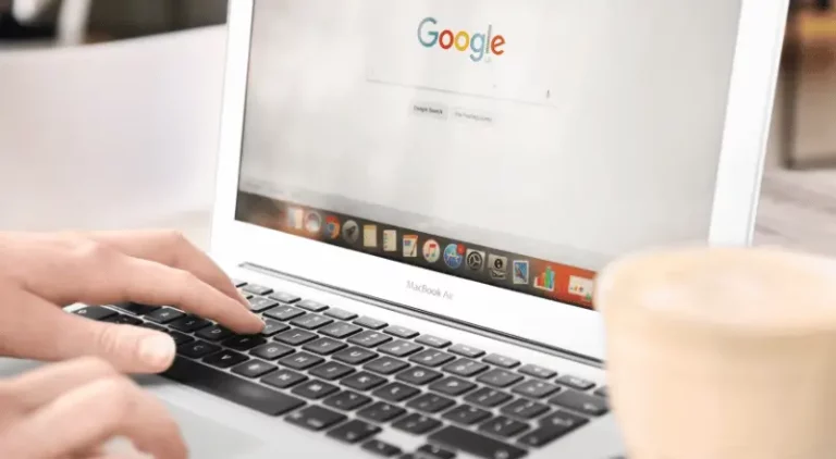 Imagem de uma pessoa navegando pela página principal do Google, em um notebook. Aparece só as mãos dessa pessoa e há um copo de café ao lado da tela do computador. A pessoa é branca.